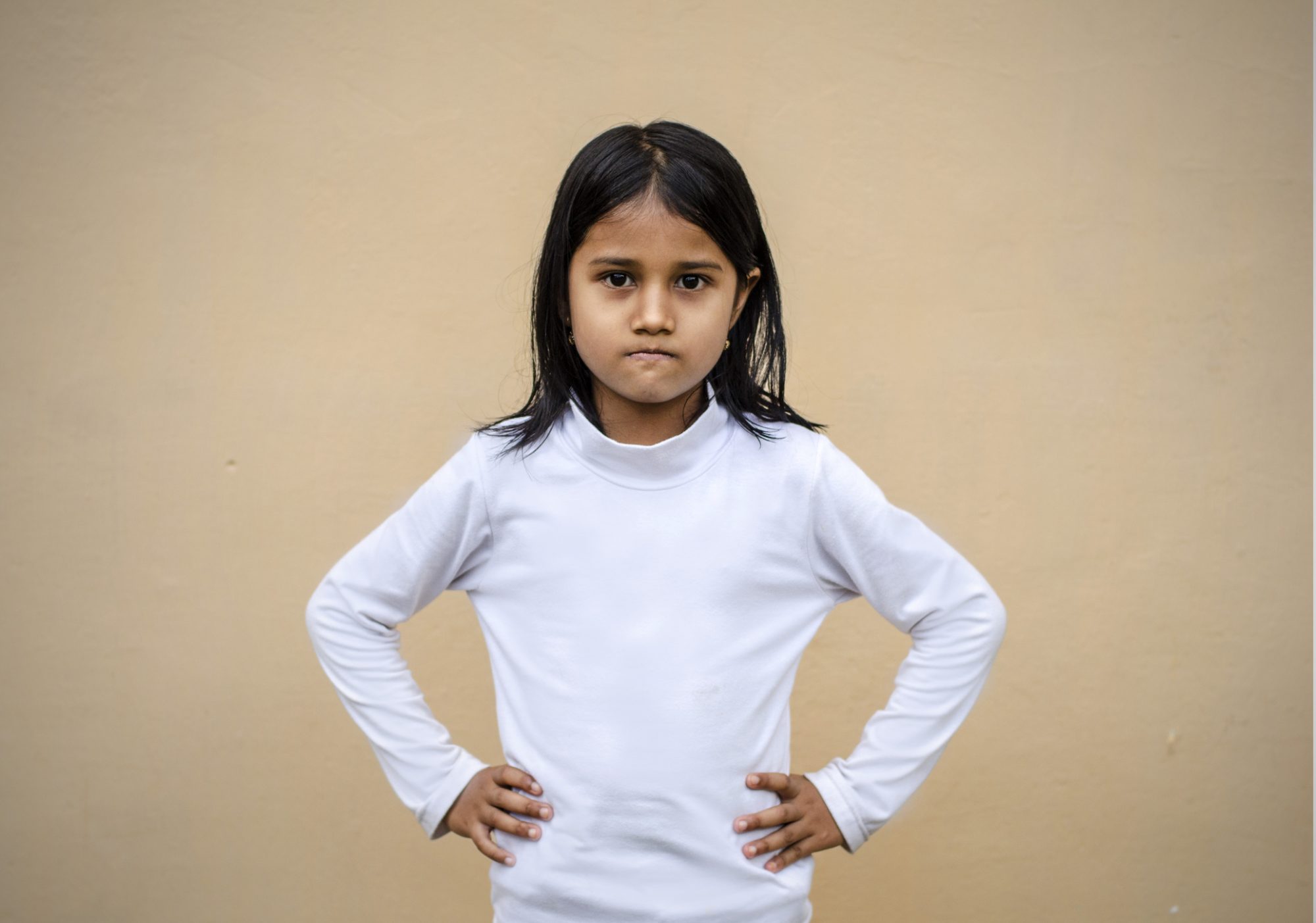 girl with stern expression standing against blank beige wall with arms on hips